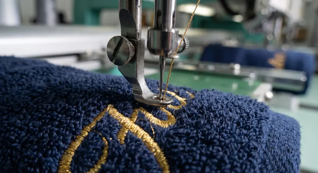 Industrial embroidery machine stitching corporate logo onto thick navy blue hotel towel in textile factory